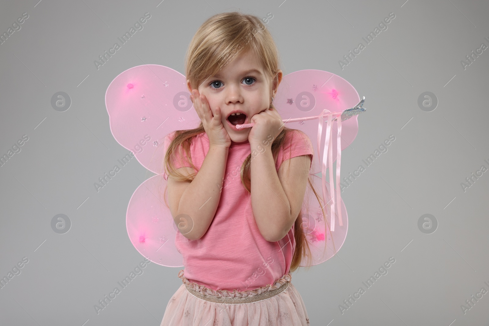 Photo of Surprised little girl in fairy costume with pink wings and magic wand on light grey background