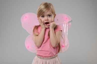 Surprised little girl in fairy costume with pink wings and magic wand on light grey background