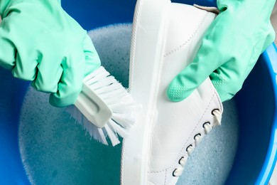 Photo of Woman with gloves and brush cleaning stylish sneakers in wash basin, top view