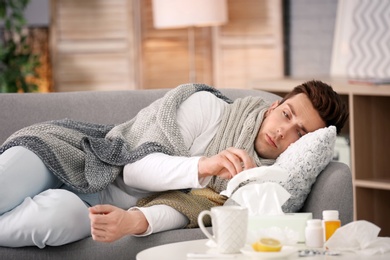 Sick young man with tissue suffering from cold on sofa at home