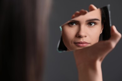 Photo of Young woman looking at herself in shard of broken mirror on grey background, closeup