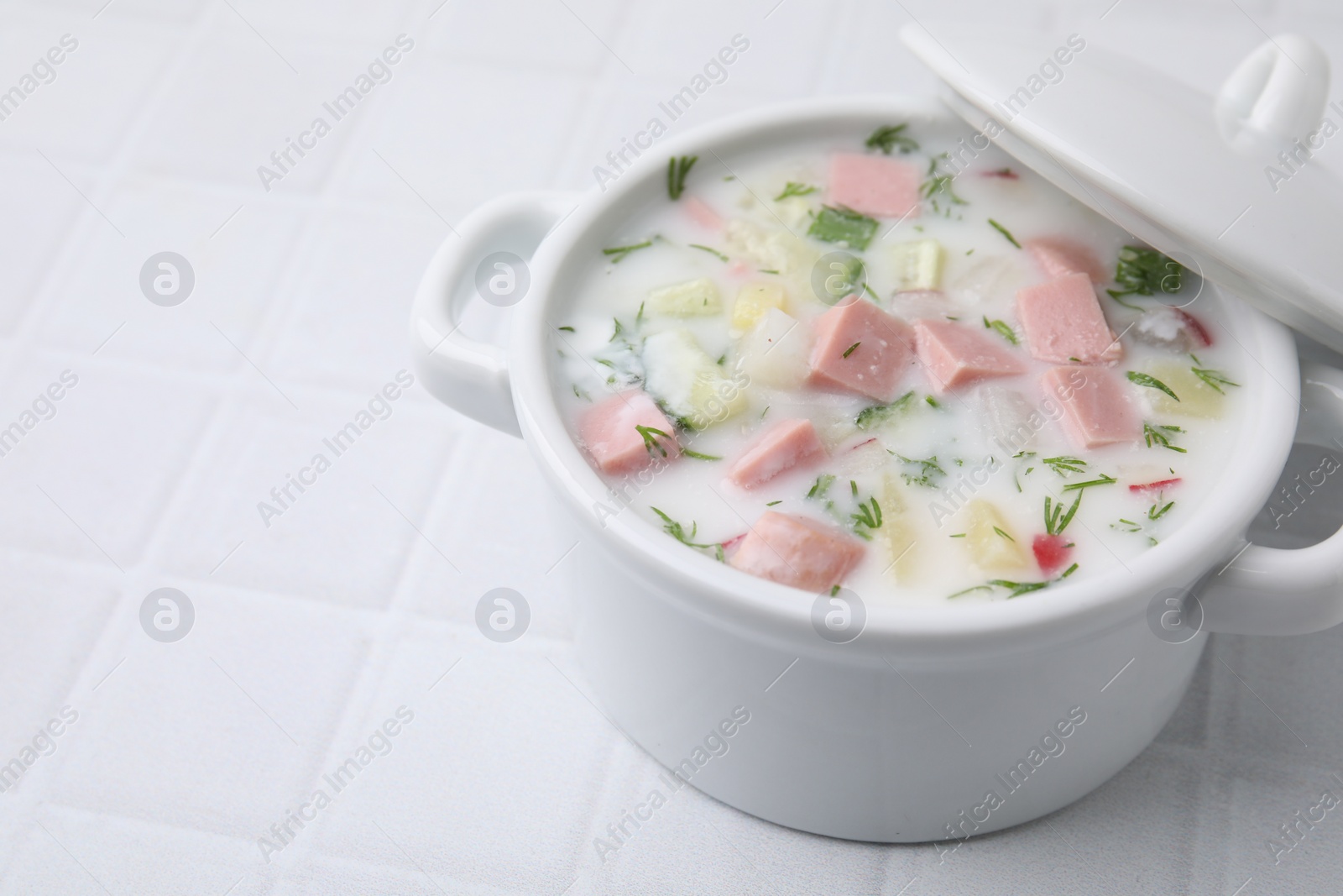 Photo of Delicious cold summer soup (okroshka) with boiled sausage in pot on white tiled table, closeup. Space for text