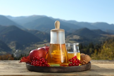 Photo of Fresh aromatic honey, rowan berries, apples and nuts on wooden table against mountain landscape