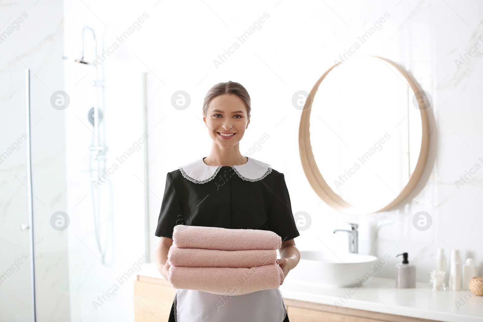 Photo of Young chambermaid holding stack of fresh towels in bathroom