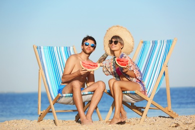 Young couple with watermelon slices in beach chairs at seacoast