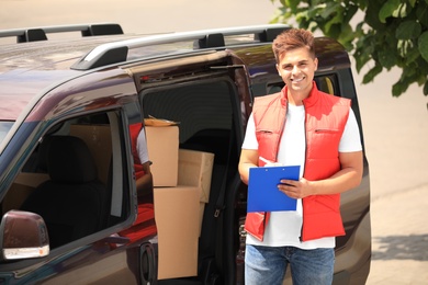 Photo of Young courier with clipboard near delivery car outdoors