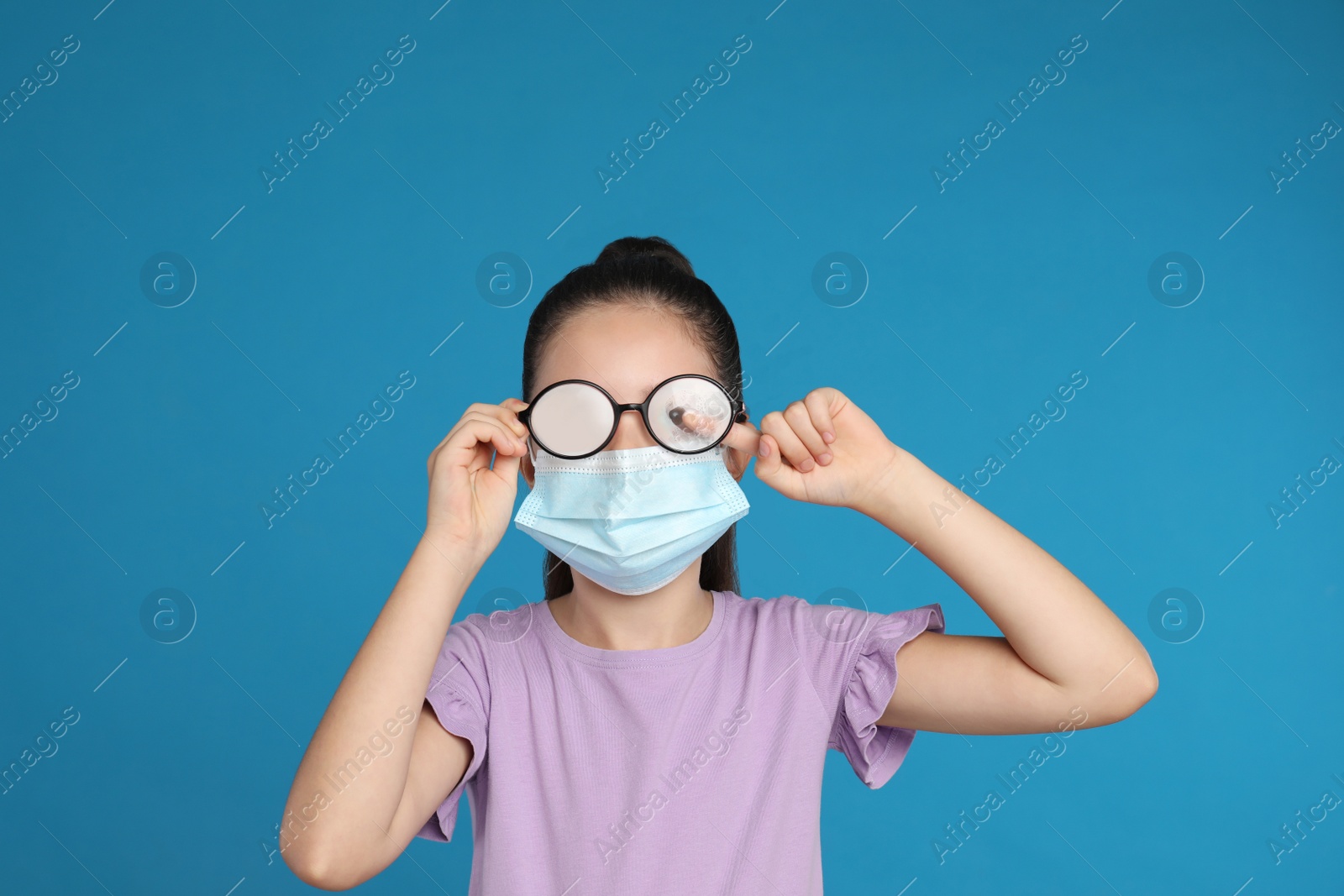 Photo of Little girl wiping foggy glasses caused by wearing medical face mask on blue background. Protective measure during coronavirus pandemic