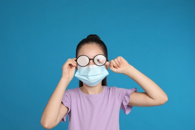 Photo of Little girl wiping foggy glasses caused by wearing medical face mask on blue background. Protective measure during coronavirus pandemic