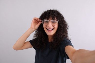 Beautiful young woman in eyeglasses taking selfie on light grey background