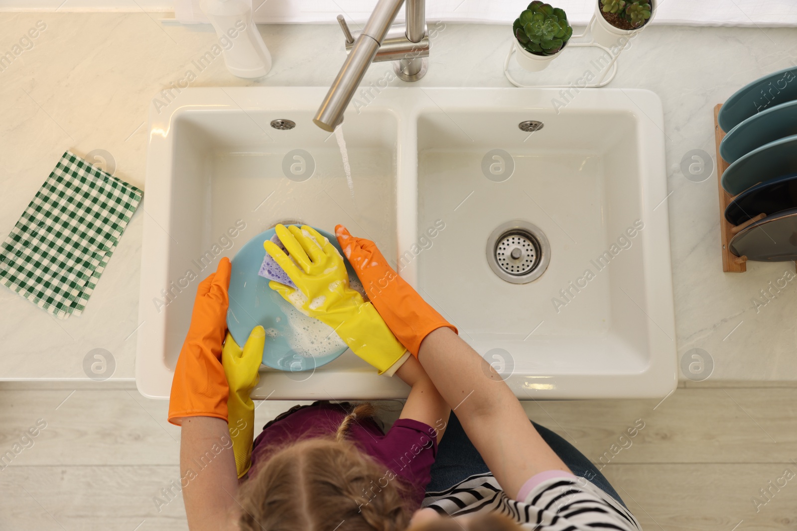 Photo of Mother and daughter in protective gloves washing plate above sink, top view