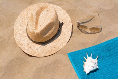 Soft blue beach towel, sunglasses, straw hat and seashell on sand, flat lay