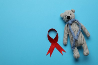 Cute knitted toy bear and red ribbon on blue background, flat lay with space for text. AIDS disease awareness
