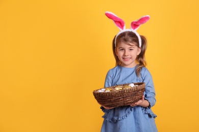 Photo of Happy little girl with bunny ears holding wicker basket full of Easter eggs on orange background. Space for text