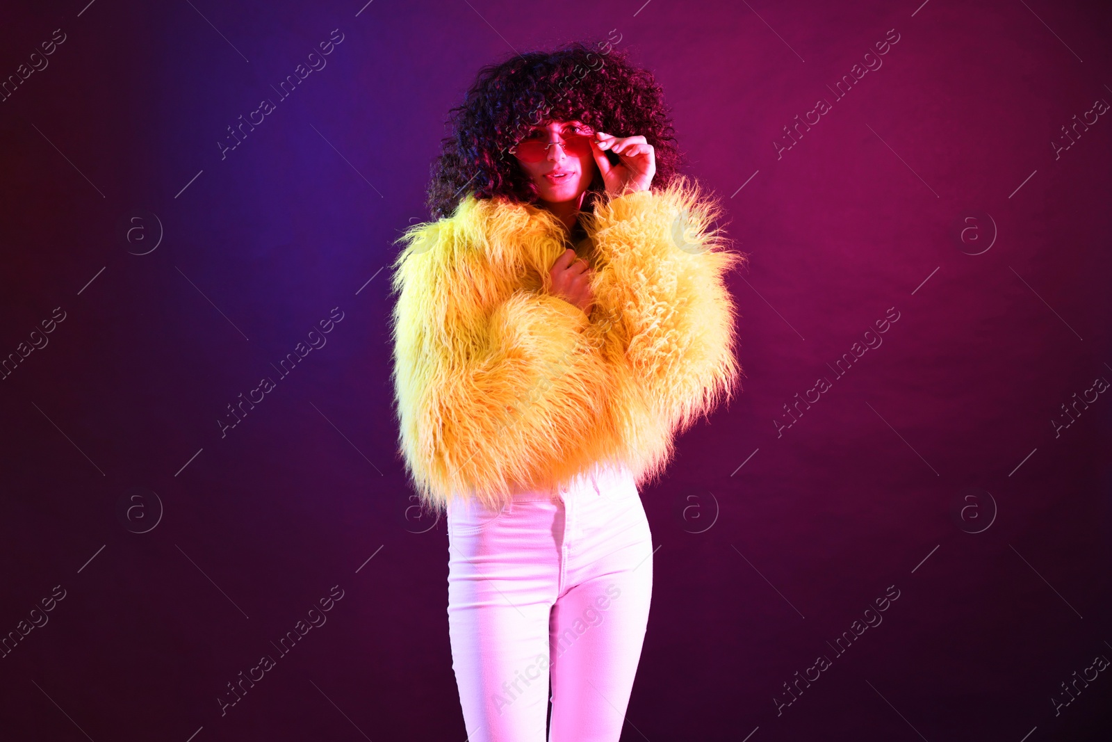 Photo of Beautiful young woman in yellow fur coat and sunglasses on color background in neon lights