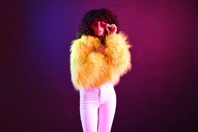 Photo of Beautiful young woman in yellow fur coat and sunglasses on color background in neon lights