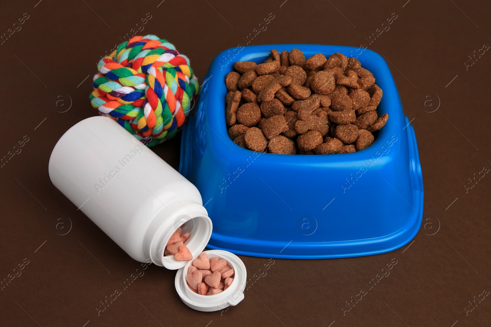 Photo of Bowl with dry pet food, bottle of vitamins and toy on brown background
