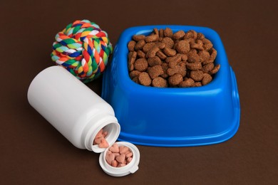 Bowl with dry pet food, bottle of vitamins and toy on brown background