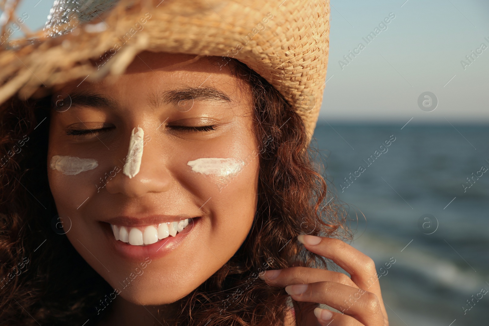 Photo of Happy African American woman with sun protection cream on face near sea, closeup