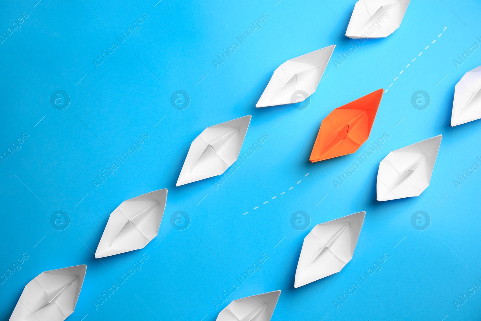 Photo of Orange paper boat floating between others on light blue background, flat lay. Uniqueness concept