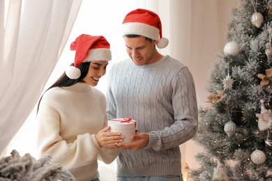 Photo of Couple in Santa hats holding gift box near Christmas tree