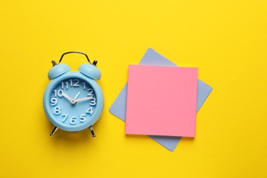 Alarm clock and blank reminder notes on yellow background, flat lay