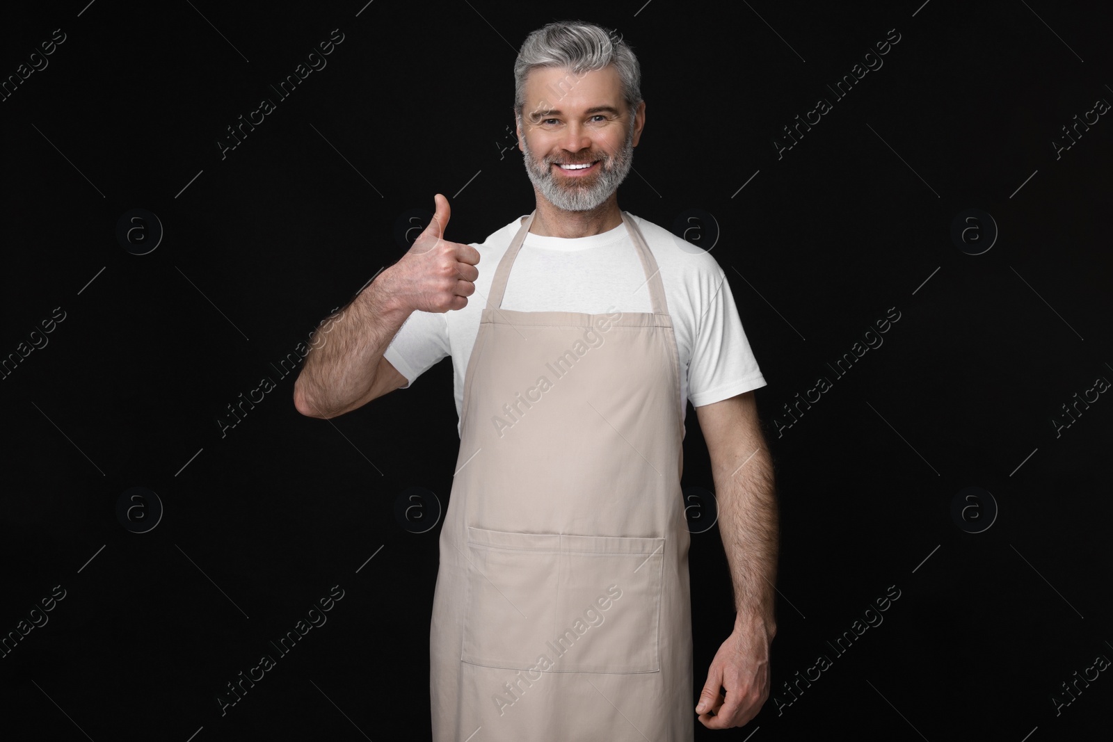 Photo of Happy man in kitchen apron showing thumbs up on black background. Mockup for design