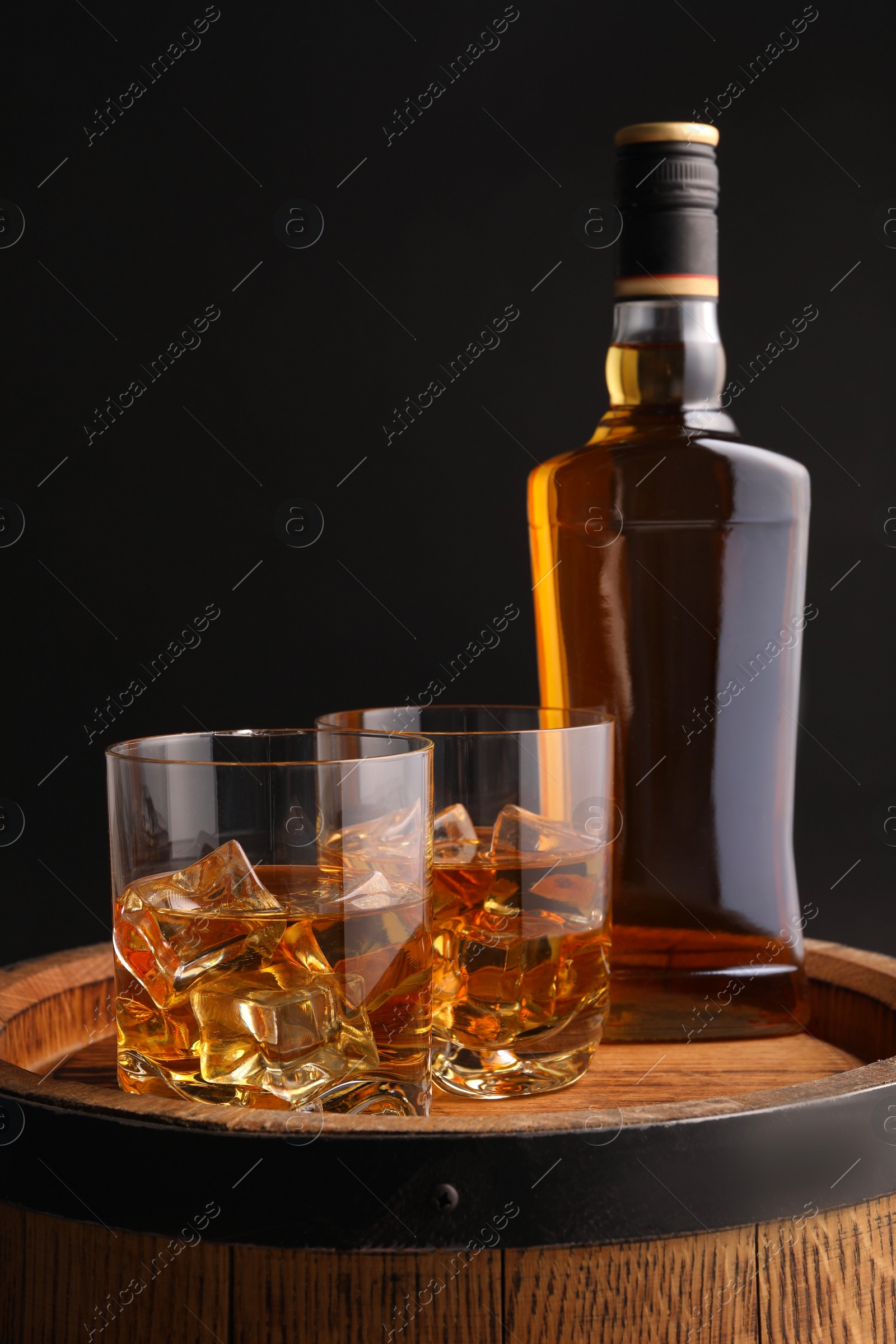 Photo of Whiskey with ice cubes in glasses and bottle on wooden barrel against black background