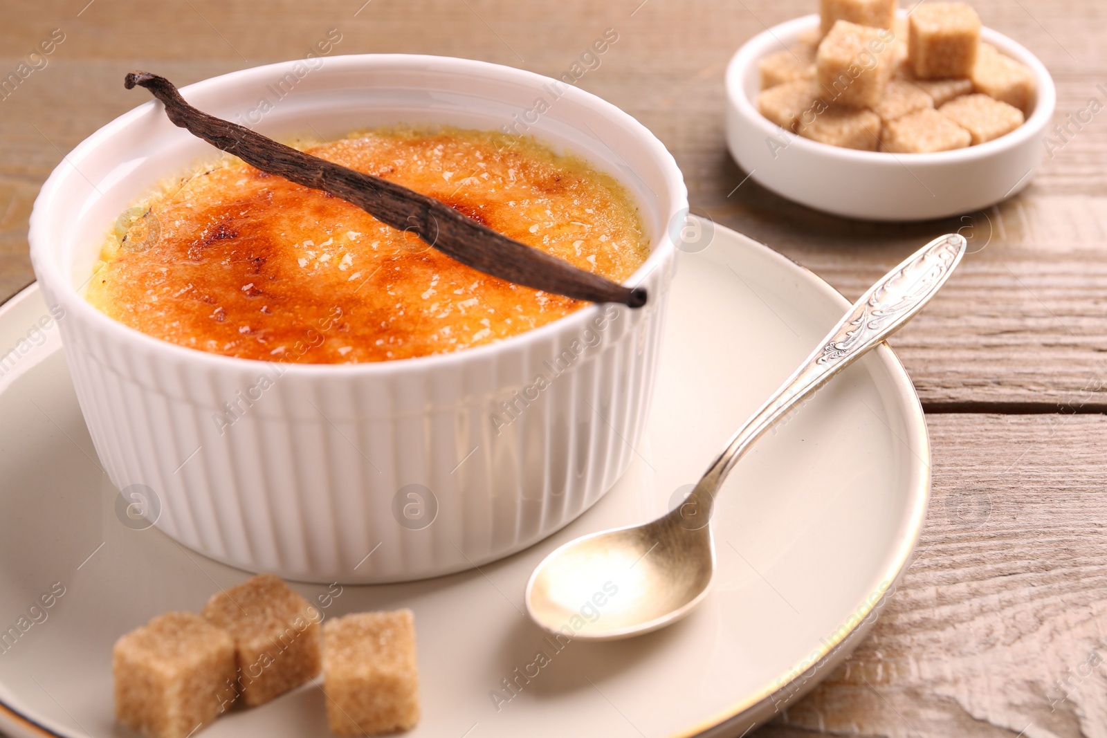 Photo of Delicious creme brulee in bowl, vanilla pod, spoon and sugar cubes on wooden table, closeup