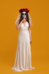 Young woman in scary bride costume with sugar skull makeup and flower crown showing shush gesture on orange background. Halloween celebration