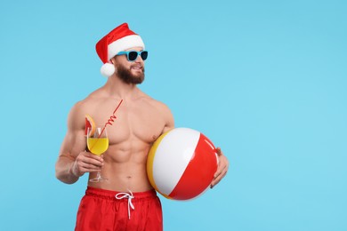 Photo of Attractive young man with muscular body in Santa hat holding ball and cocktail on light blue background, space for text