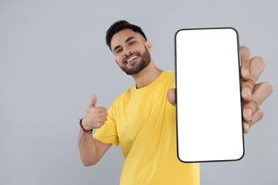 Happy man holding smartphone with empty screen and showing thumbs up on grey background, space for text