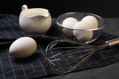 Photo of Metal whisk, raw eggs and milk on dark grey table, closeup