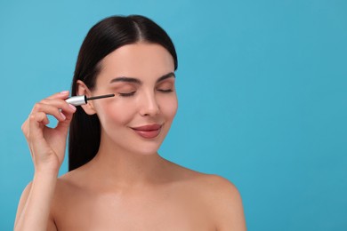 Photo of Beautiful woman applying serum onto her eyelashes on light blue background, space for text. Cosmetic product