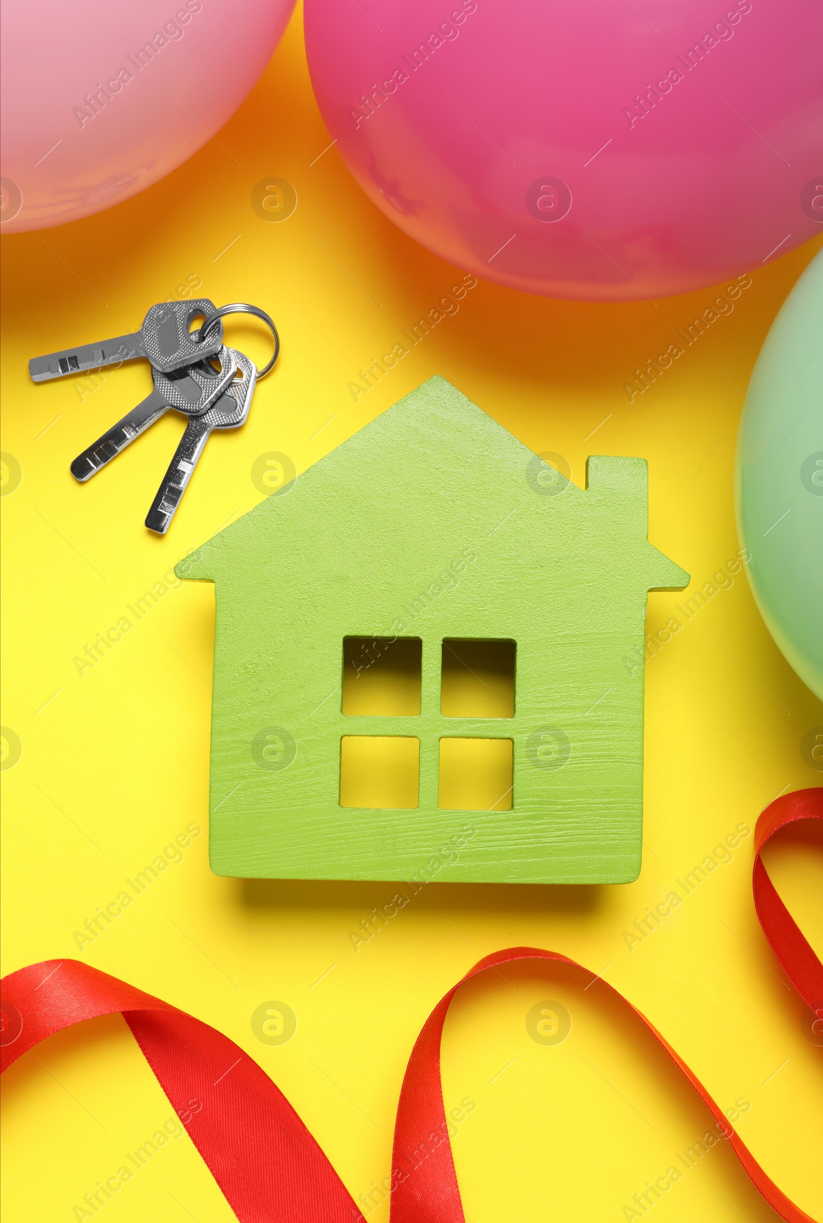 Photo of Wooden house model, keys, red ribbon and balloons on yellow background, flat lay. Housewarming party