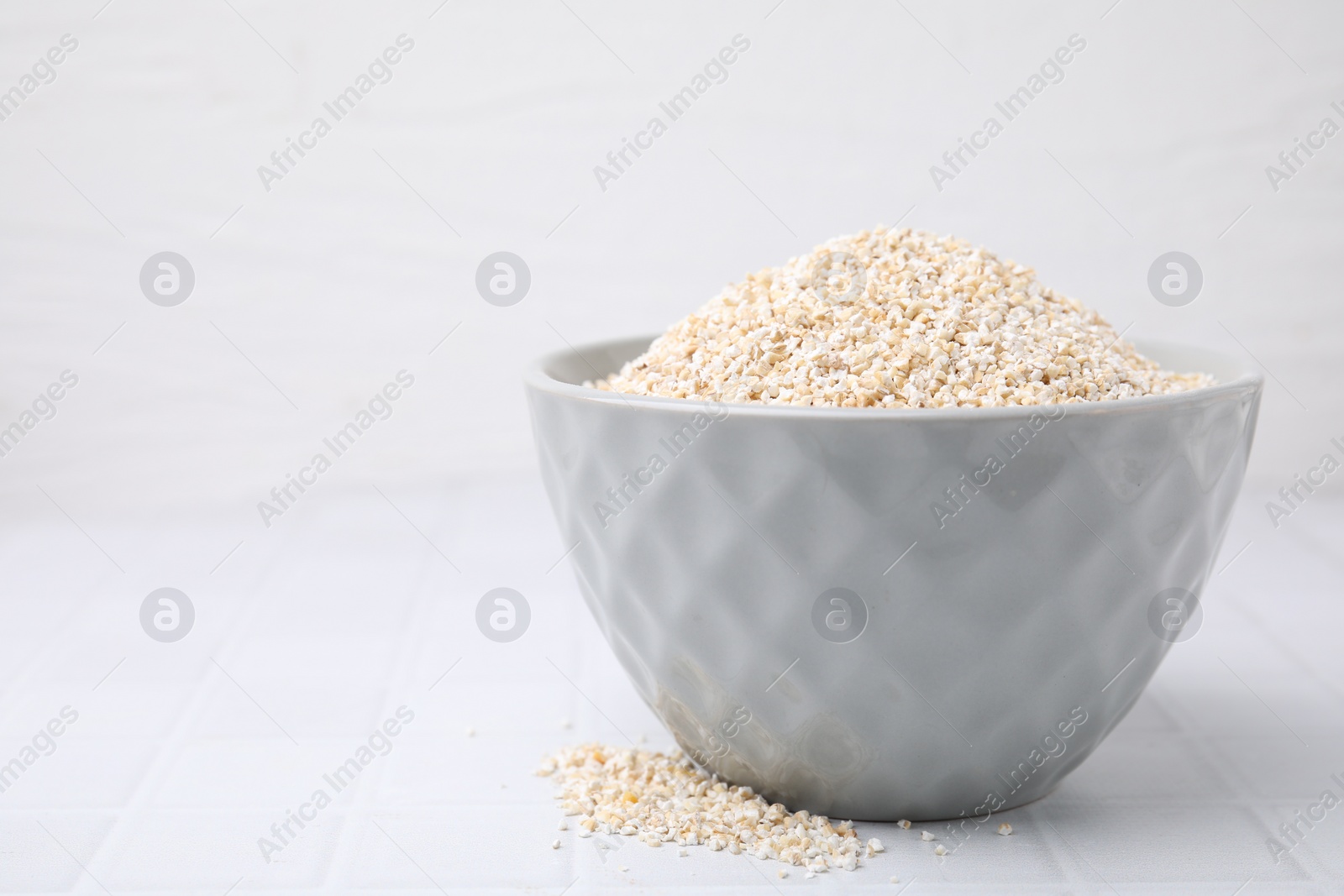 Photo of Dry barley groats in bowl on white tiled table, closeup. Space for text
