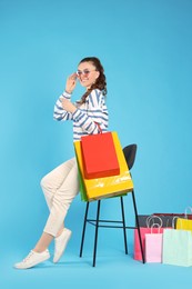 Happy woman in stylish sunglasses holding colorful shopping bags on stool against light blue background