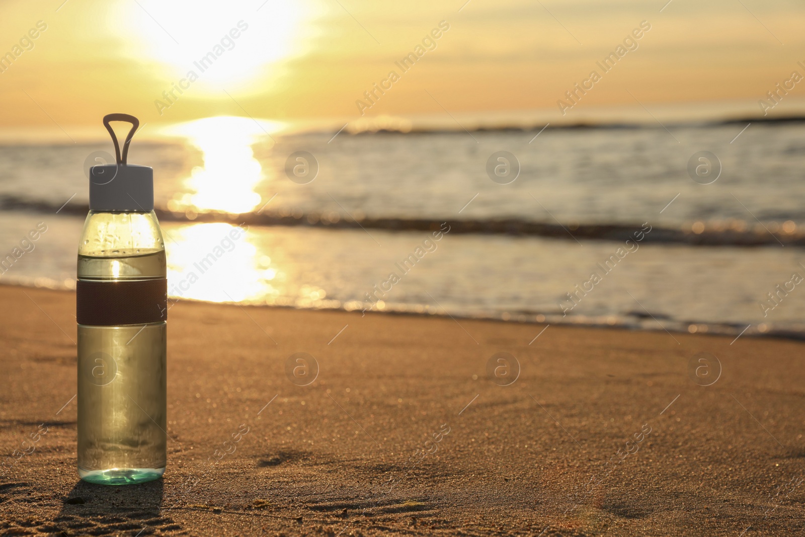 Photo of Glass bottle with water on wet sand near sea at sunset. Space for text