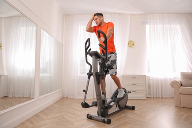 Tired man using modern elliptical machine at home