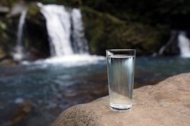 Glass of water on stone near waterfall outdoors. Space for text