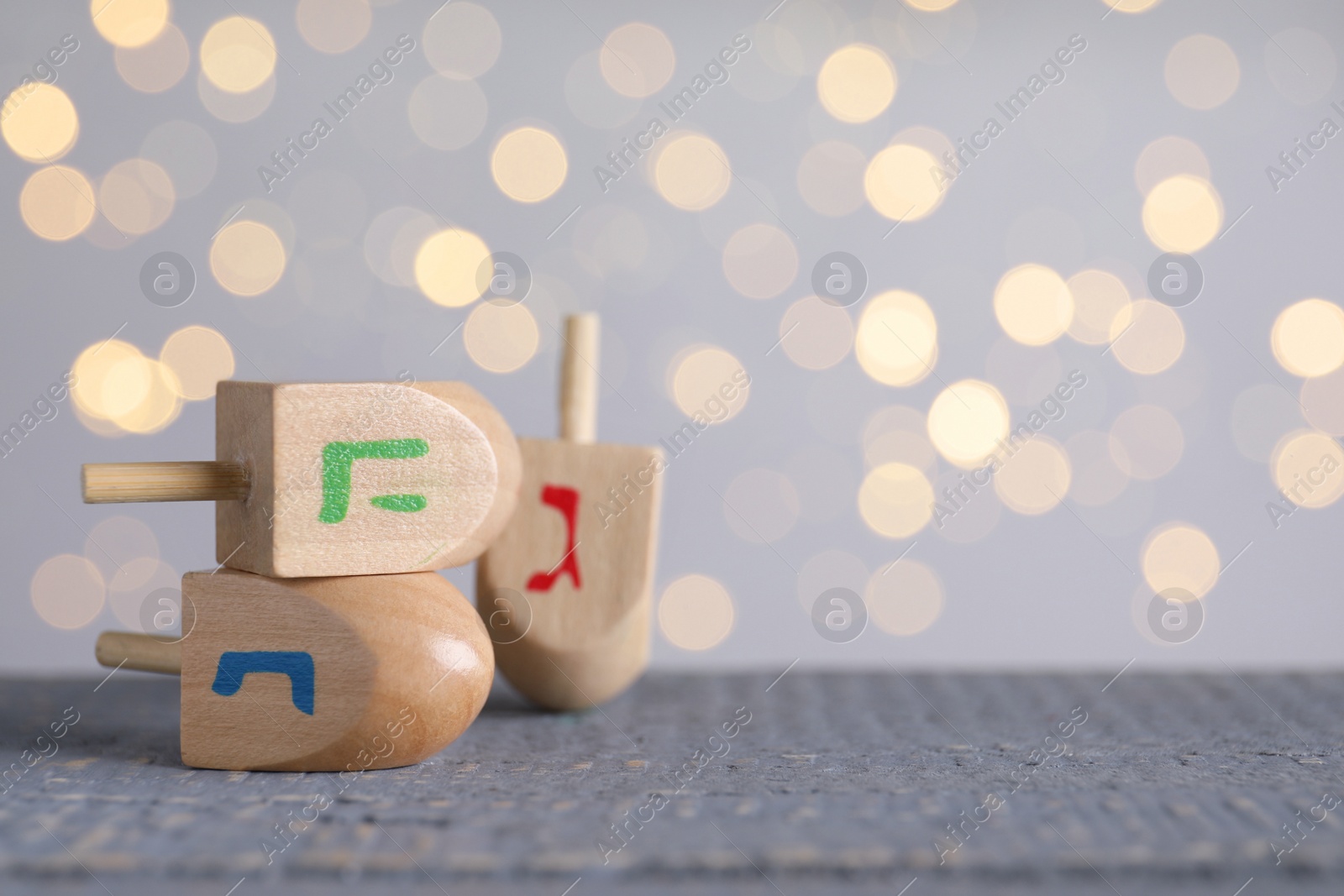 Photo of Hanukkah traditional dreidels with letters Nun, He and Gimel on grey wooden table against blurred lights. Space for text