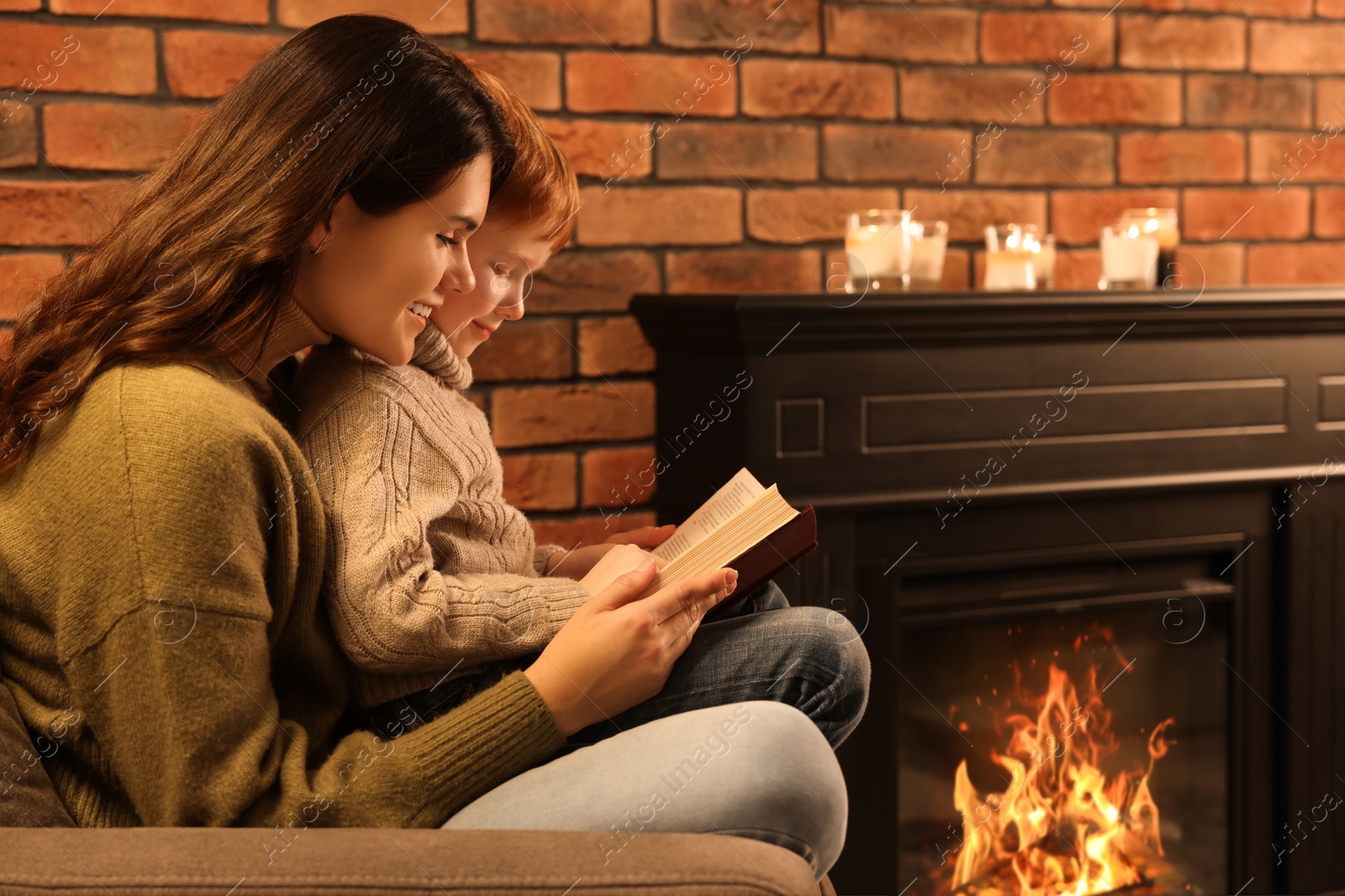 Photo of Happy mother and son reading book together near fireplace at home