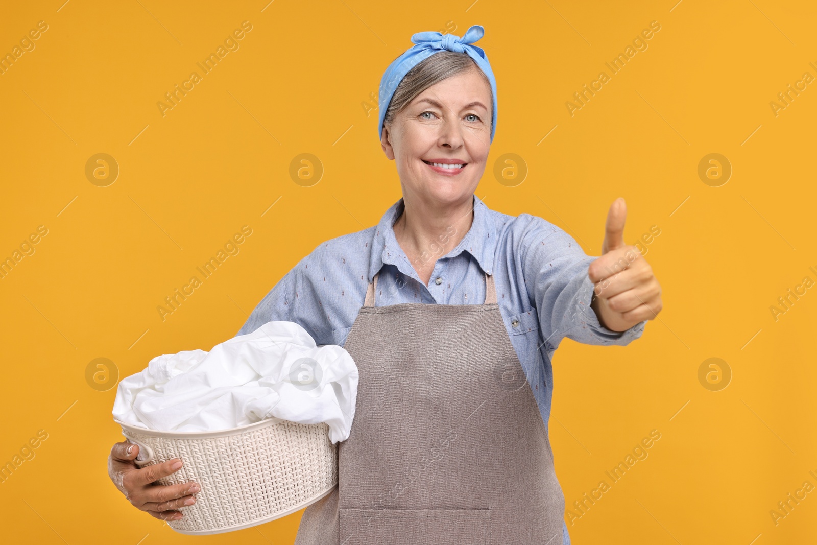 Photo of Happy housewife with basket full of laundry showing thumbs up on orange background