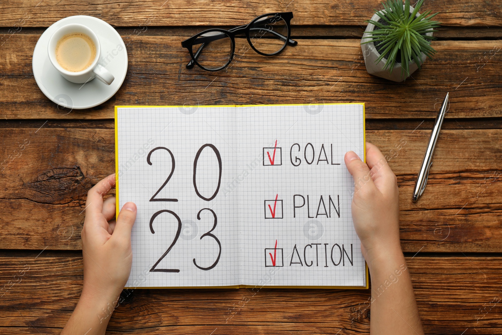Photo of Woman holding notebook with text 2023 Goal, Plan, Action at wooden table, top view. New Year resolutions