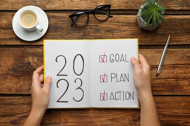 Photo of Woman holding notebook with text 2023 Goal, Plan, Action at wooden table, top view. New Year resolutions