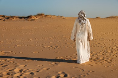 Man in arabic clothes walking through desert on sunny day, back view