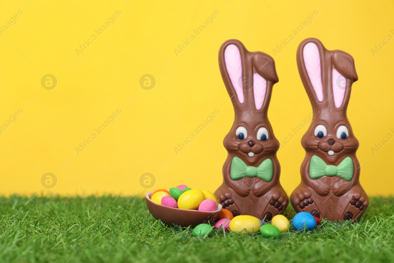 Photo of Easter celebration. Funny chocolate bunnies and half of sweet egg with small candies on grass against yellow background. Space for text