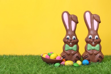 Photo of Easter celebration. Funny chocolate bunnies and half of sweet egg with small candies on grass against yellow background. Space for text