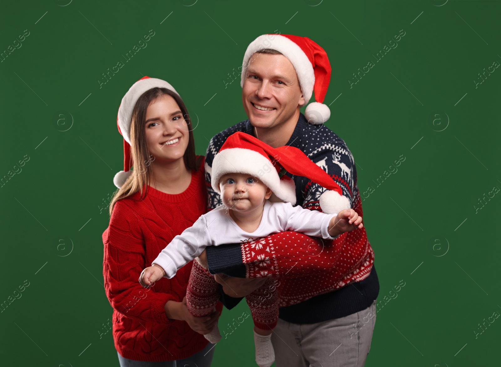 Photo of Happy couple with cute baby in Christmas outfits and Santa hats on green background