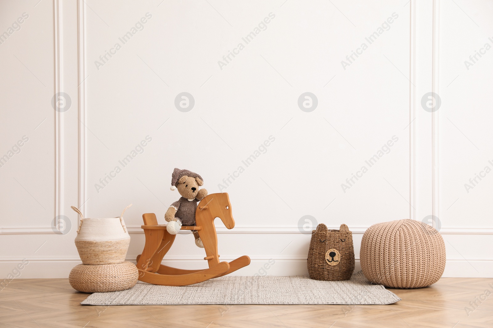 Photo of Rocking horse with bear toy, pouf and wicker baskets near white wall in child room. Interior design