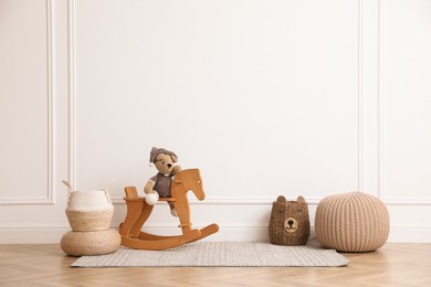 Photo of Rocking horse with bear toy, pouf and wicker baskets near white wall in child room. Interior design